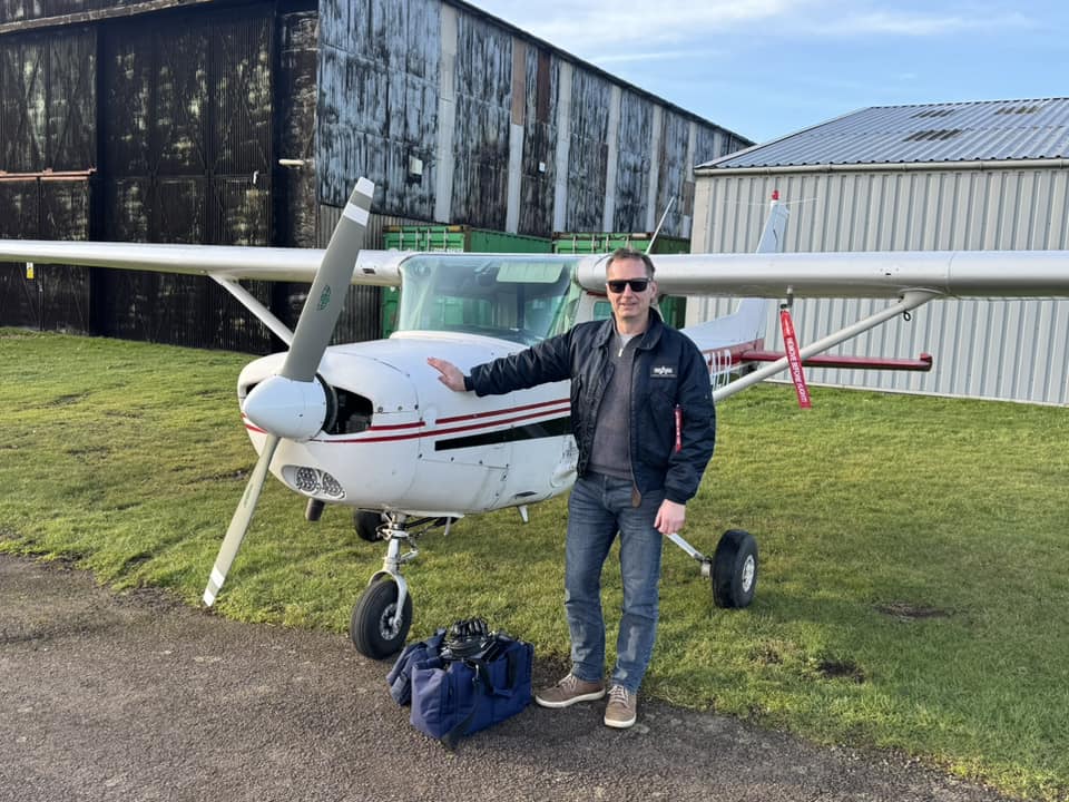 Pilot stood next to light aircraft parked on some grass outside a hangar.